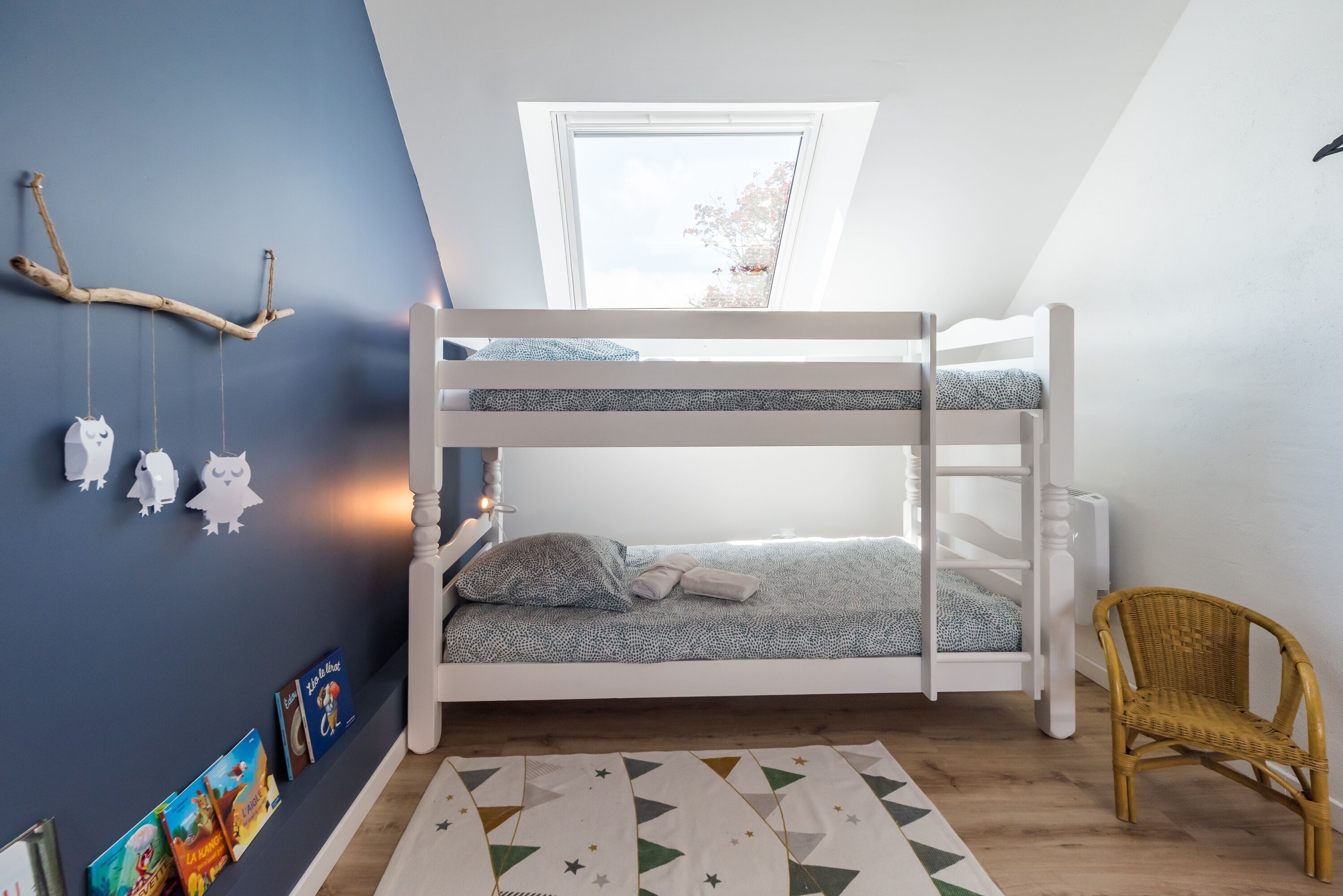 A bunk bed with a window above, a rattan chair, and a geometric rug.