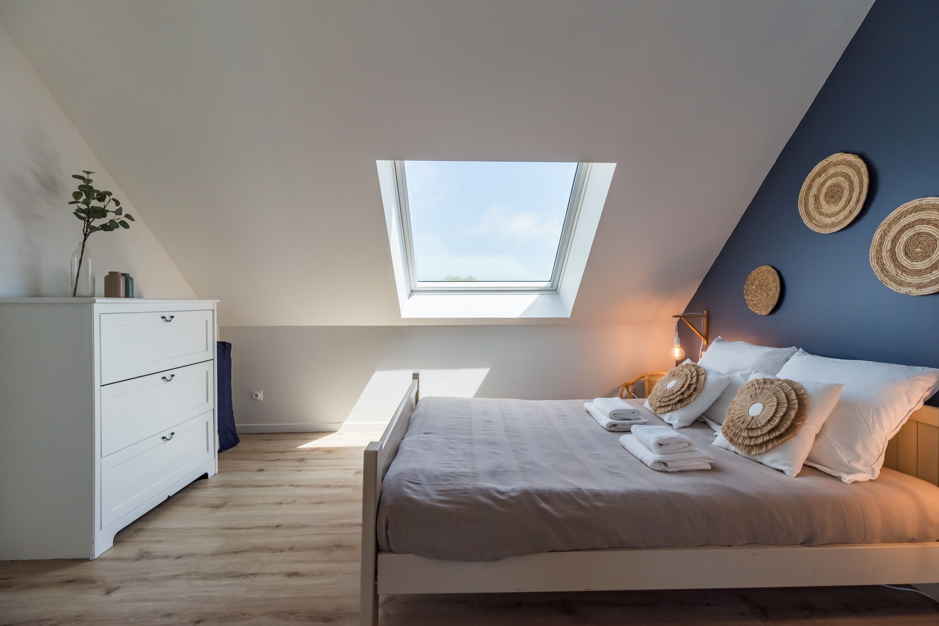 A bedroom with a skylight, a bed with pillows, a dresser, and wall decor.