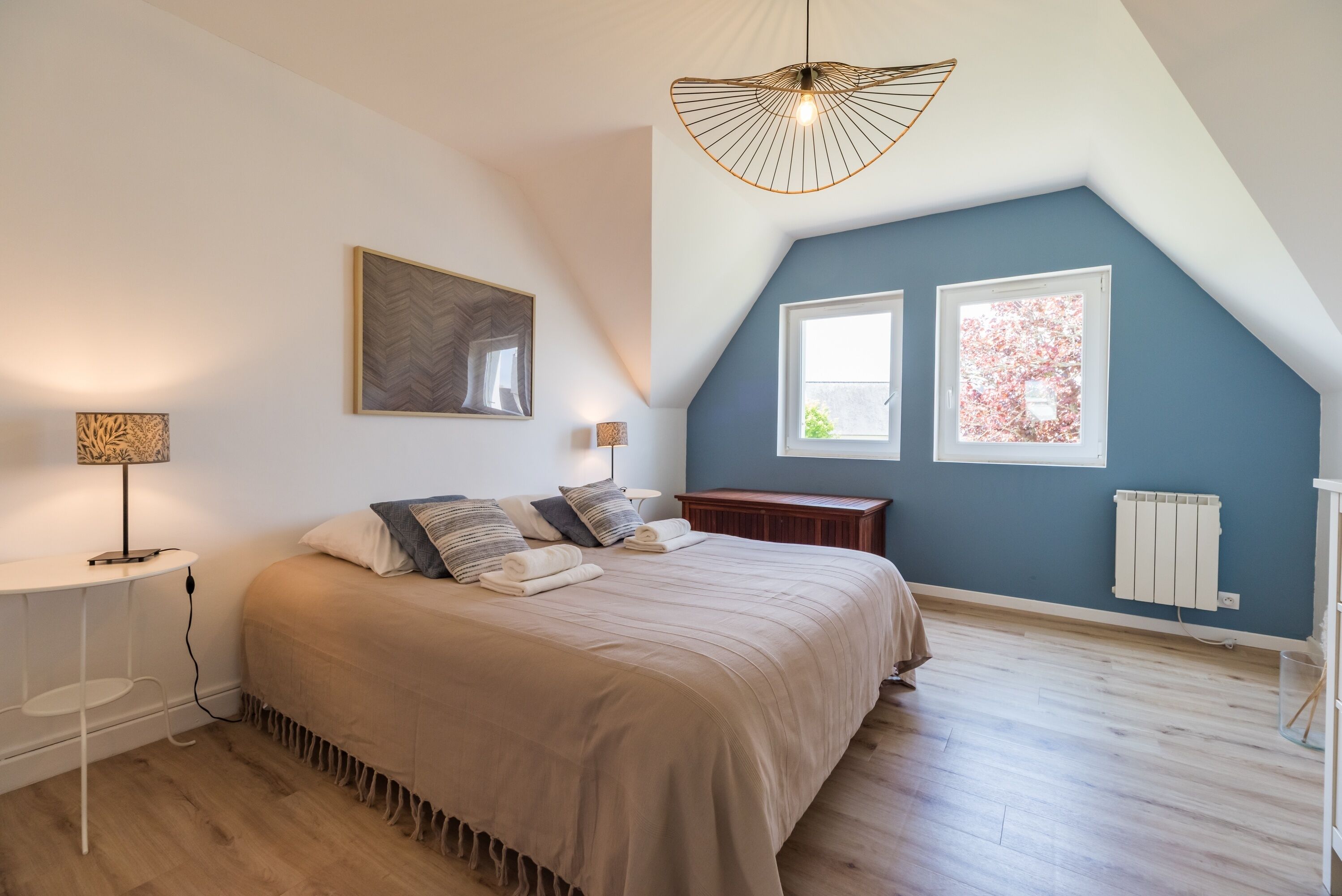 Une chambre à coucher comprenant un lit, des tables de chevet, un luminaire suspendu et deux fenêtres.