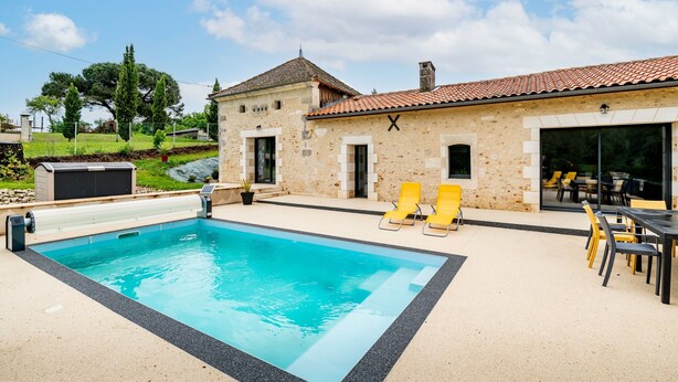 Une piscine avec un bâtiment en pierre en arrière-plan, des sièges d’extérieur et un ciel dégagé.