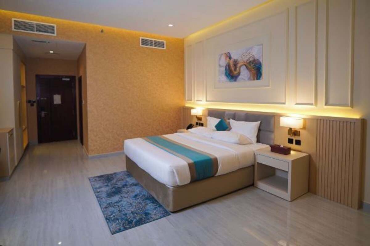 A hotel room with a bed, bedside tables, a nightstand, a wall-mounted artwork, and a patterned rug.