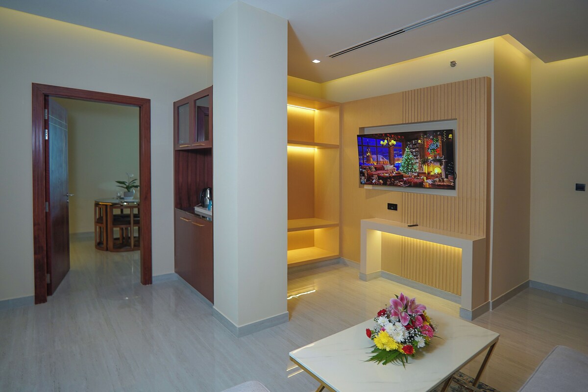 A modern living room with a flat-screen TV mounted on the wall, a dining table with a floral centerpiece, and a wooden cabinet.