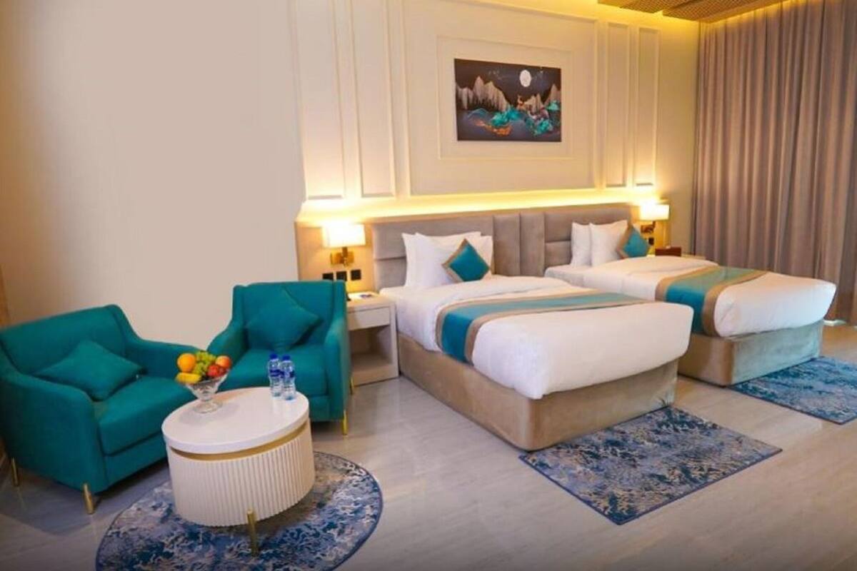 Une chambre d’hôtel avec deux lits, un fauteuil turquoise, une table basse ronde avec des fruits et un tableau au mur.