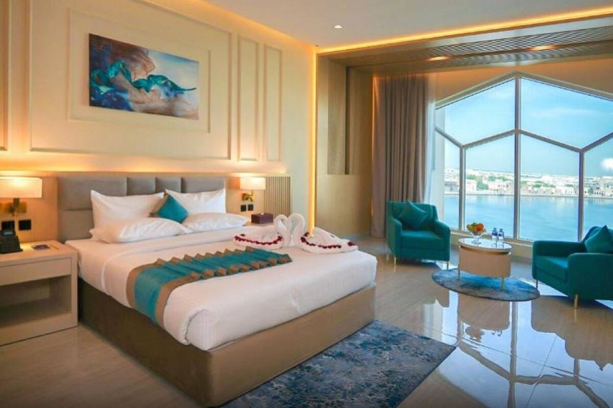 Une chambre d’hôtel moderne, dotée d’un grand lit, de deux fauteuils, d’une table basse ronde et offrant une vue sur la ville grâce à de grandes fenêtres.