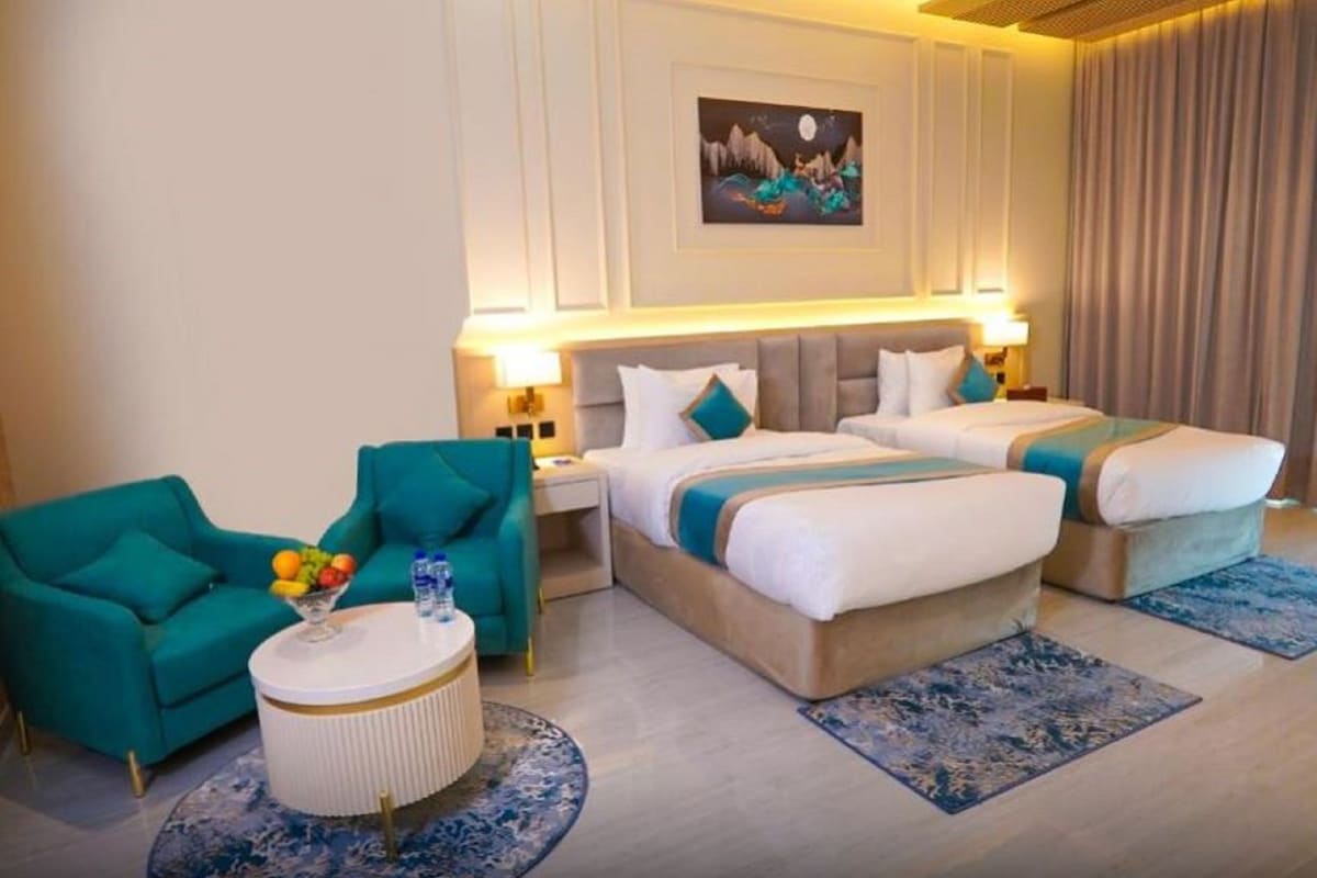 Habitación de hotel con dos camas, un sillón azul turquesa, una mesa de centro redonda con fruta y un cuadro en la pared.