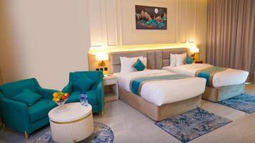 A hotel room with two beds, a teal armchair, a round coffee table with fruit, and a painting on the wall.