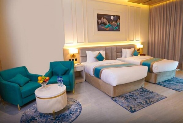 Habitación de hotel con dos camas, un sillón azul turquesa, una mesa de centro redonda con fruta y un cuadro en la pared.