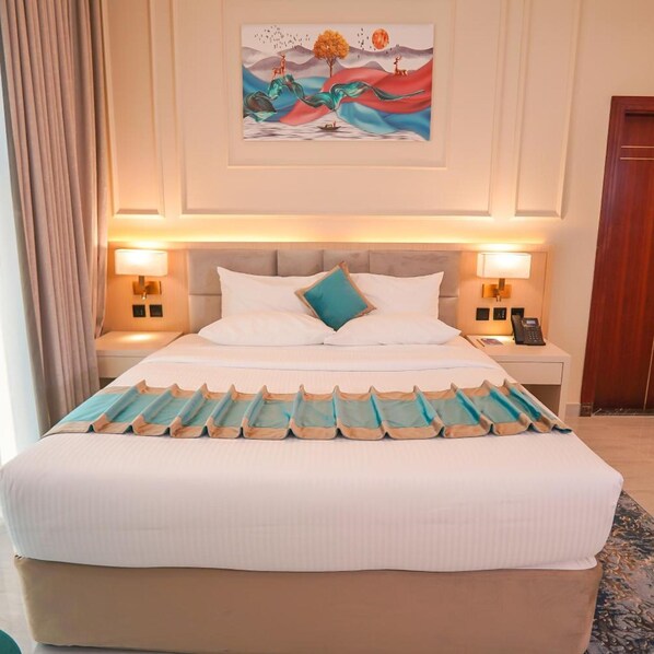 A hotel room with a large bed, two bedside tables, a nightstand, a painting on the wall, and a door. - Concorde Creek View Hotel (Dubai)