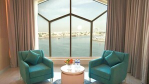 A room with two teal armchairs, a round coffee table with fruits and bottles, and a large window with a view of a bridge and water. - Concorde Creek View Hotel (Dubai)
