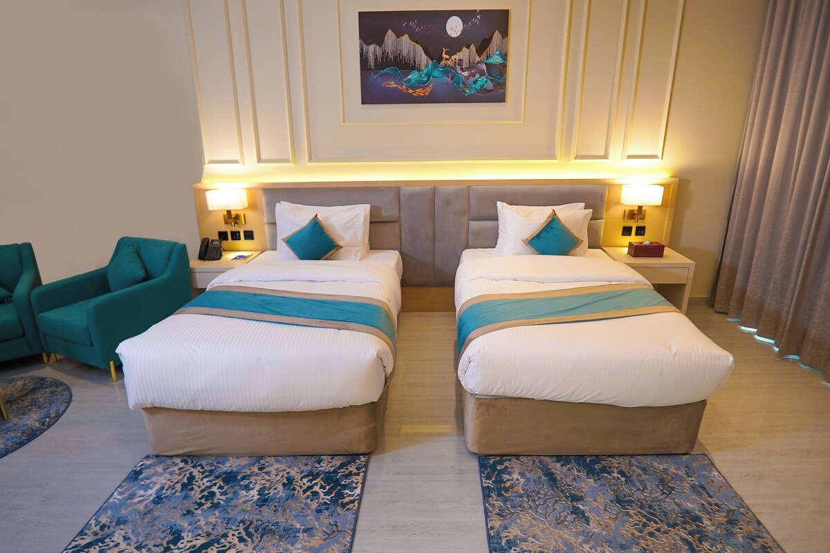 Habitación de hotel con dos camas, una mesita de noche con lámpara, un cuadro en la pared y un sillón color aguamarina.