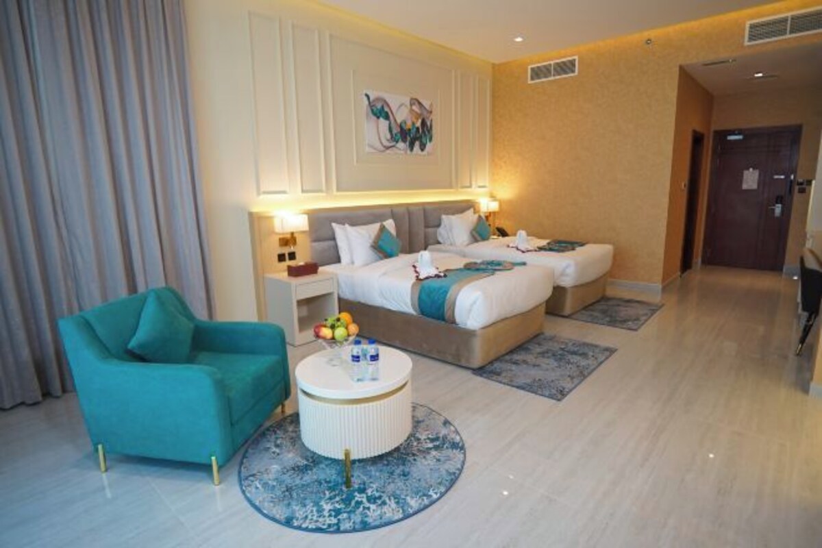 A hotel room with two beds, a teal armchair, a round coffee table, and a painting on the wall.