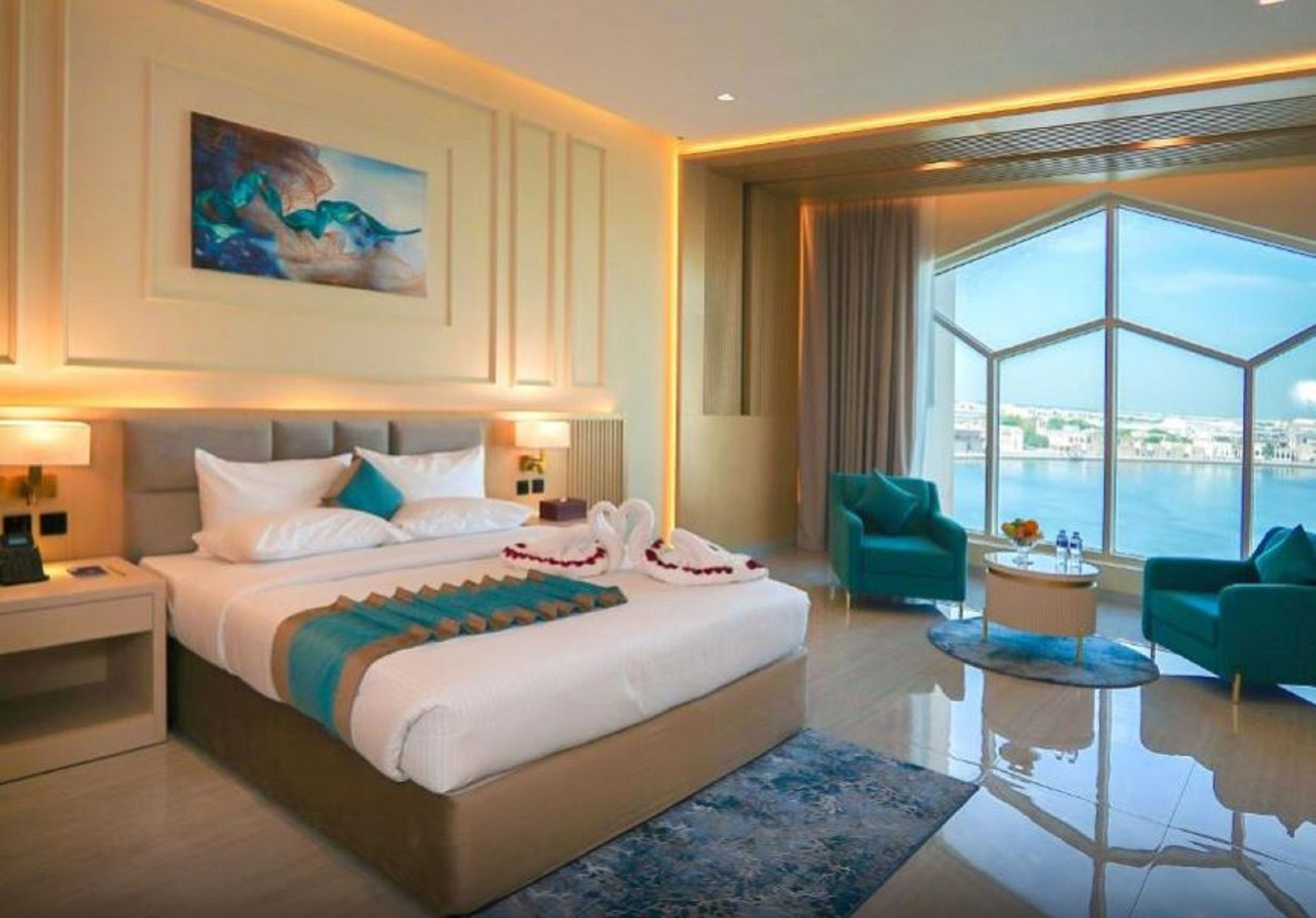A modern hotel room with a large bed, two armchairs, a round coffee table, and a view of the city through large windows.