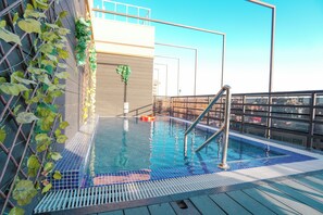 A rooftop pool with a metal ladder, a wooden deck, and a view of the cityscape.