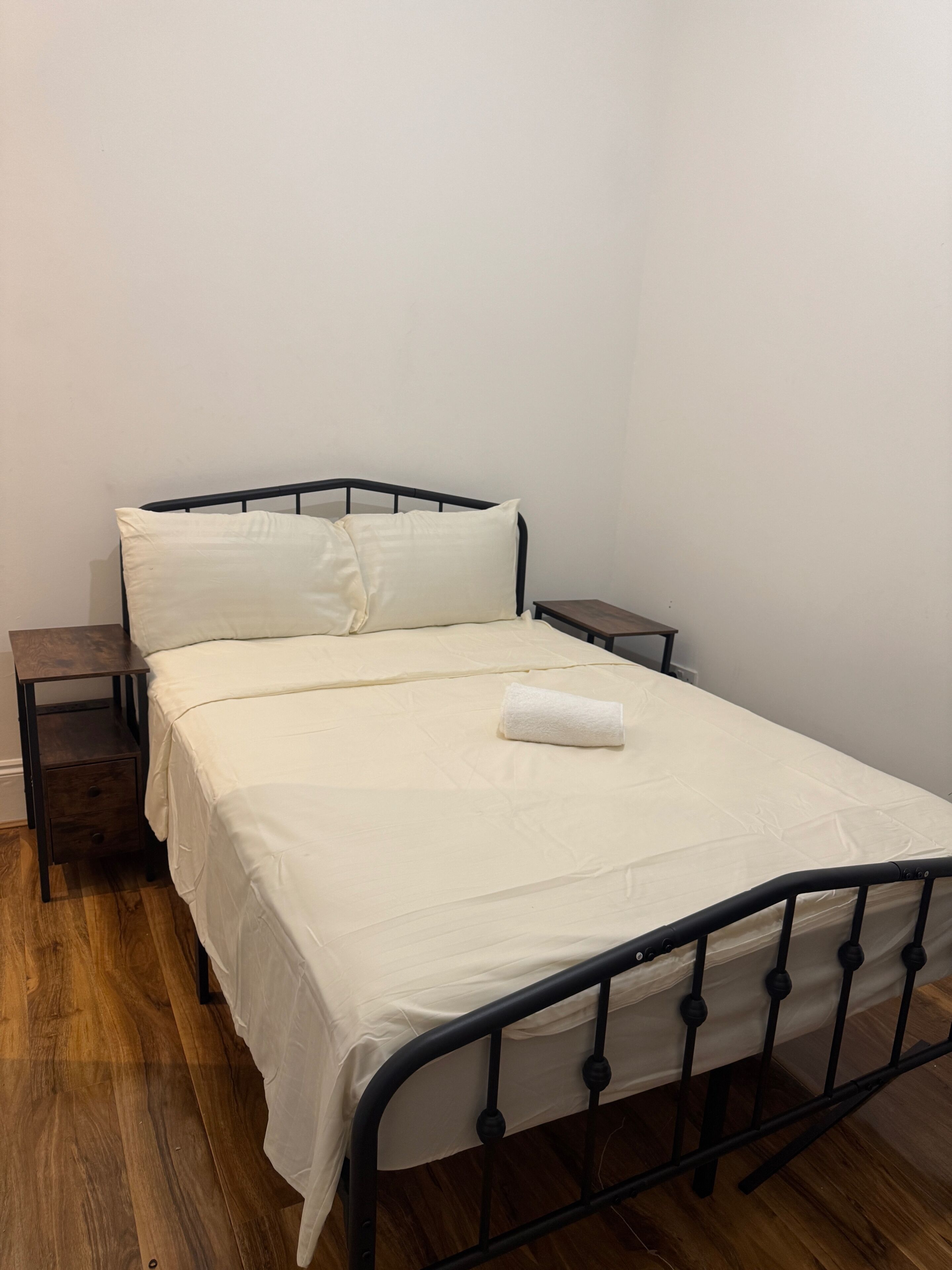 A single bed with a white mattress and matching pillows, a wooden nightstand, and a small drawer unit.