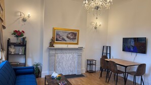 A living room with a blue sofa, a wooden coffee table, a fireplace, a chandelier, and a dining area with chairs and a TV. - 2 Bed Flat - Sleeps 4 - Parking - Wifi (London)