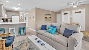 A modern living room with a beige sofa, a coffee table, and a dining area with a white counter and bar stools.