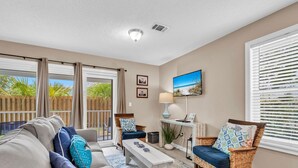 A living room with a sofa, chairs, a coffee table, and a TV. There is a sliding glass door leading to a balcony with a wooden fence and plants.