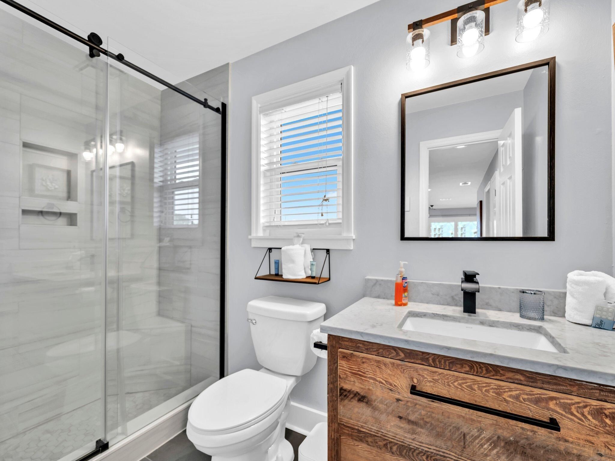 A modern bathroom with a glass shower enclosure, a toilet, a vanity with a marble countertop, and a large mirror.
