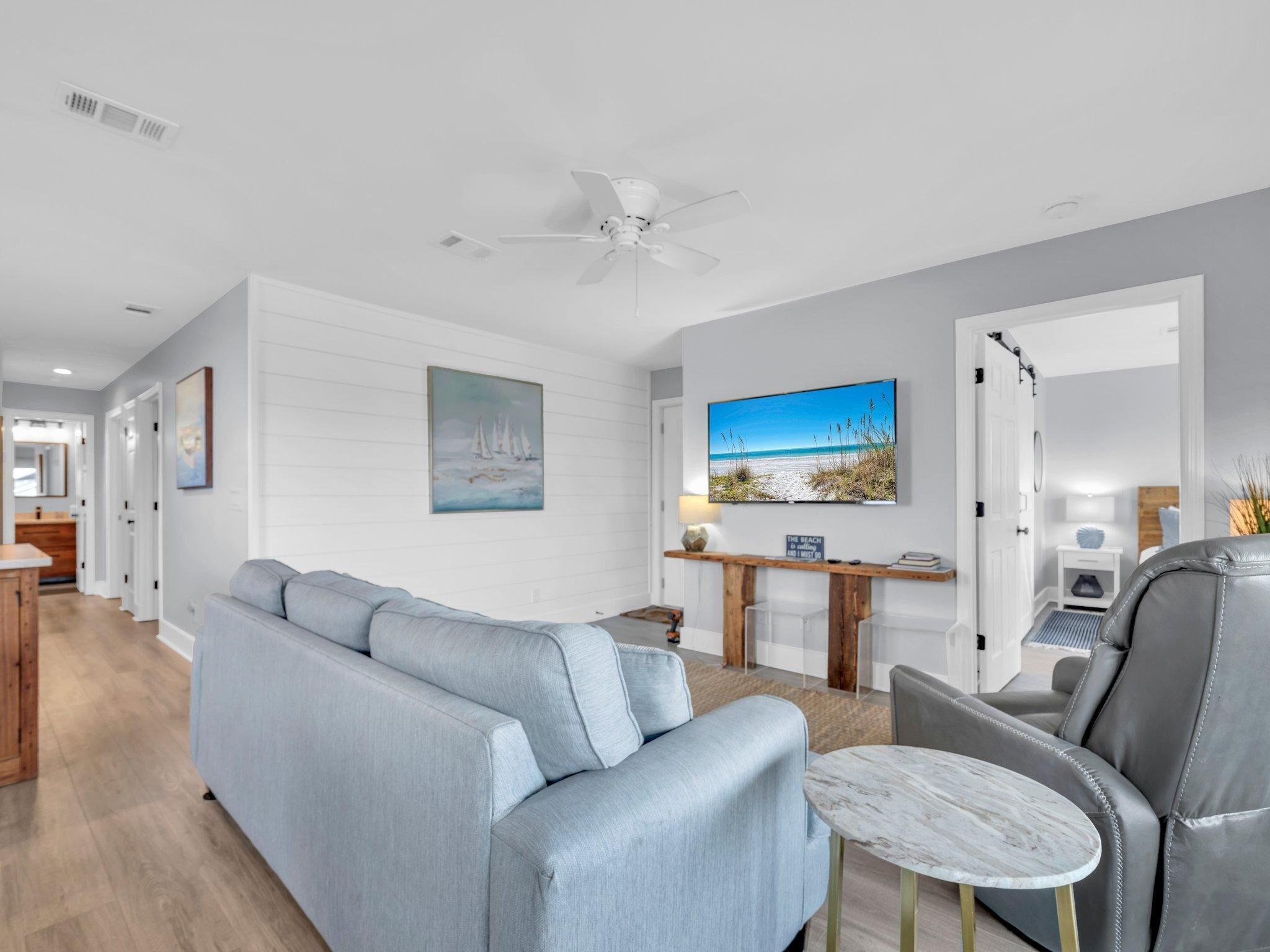 A modern living room with a light blue sofa, a grey leather chair, and a small marble table. There is a ceiling fan, a painting of a beach scene, and a wooden console table with a lamp.