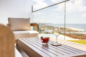 A wooden table with a glass of white wine and a bowl of strawberries on a balcony overlooking a coastal view.