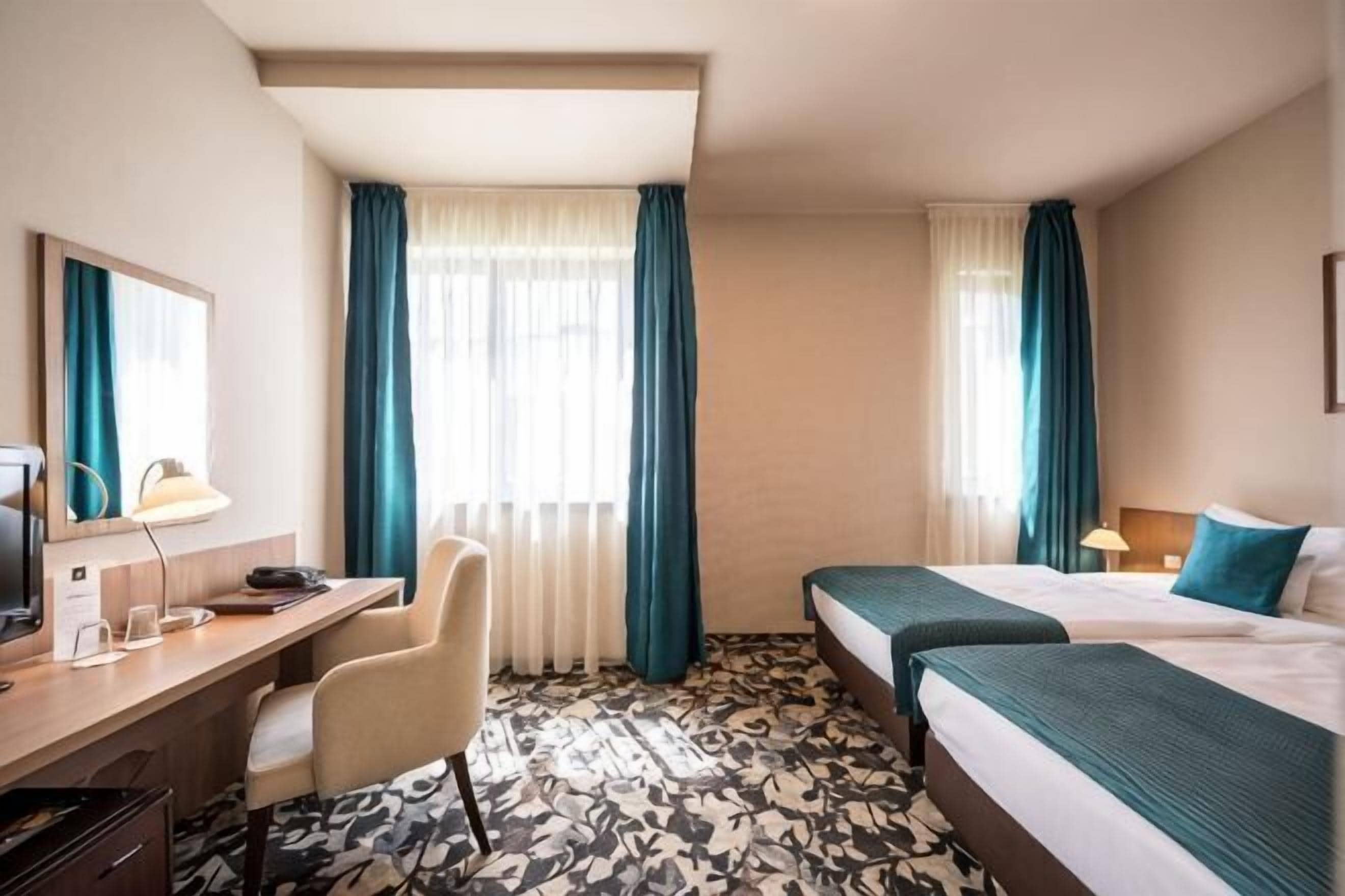 Ein Hotelzimmer mit Schreibtisch, zwei Betten, einem Sessel und einem großen Fenster mit Vorhängen.