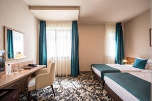 Une chambre d’hôtel équipée d’un bureau, de deux lits, d’une chaise et d’une grande fenêtre avec des rideaux.