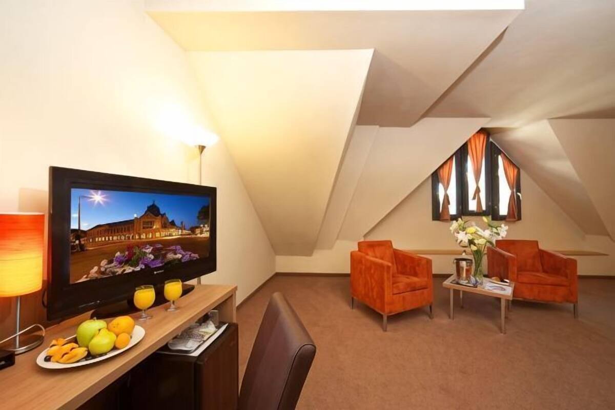 A hotel room with a television, a desk with fruit, two chairs, a small table with flowers, and a large window with curtains.