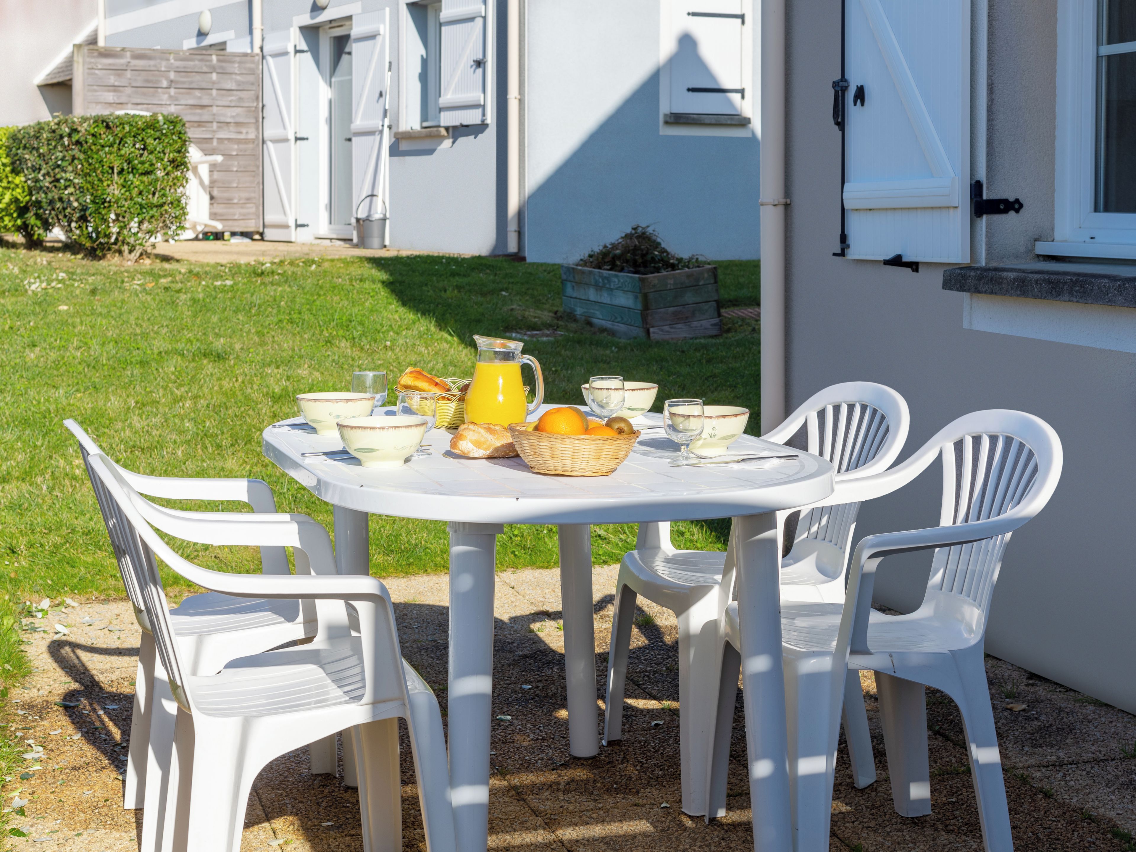 Mesa blanca al aire libre con sillas de plástico, una cesta de fruta y una jarras de zumo de naranja.