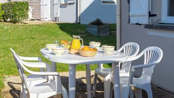 Mesa blanca al aire libre con sillas de plástico, una cesta de fruta y una jarras de zumo de naranja.