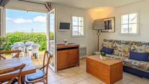 A living room with a sofa, coffee table, and dining table with chairs. There is a television and a window with a view of greenery. - Residence Vacanceole Les Terrasses De Pentrez (Saint-Nic)