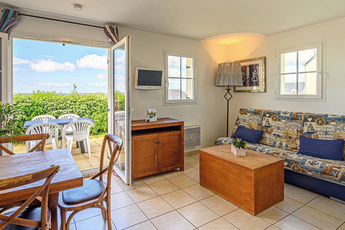 Ein Wohnzimmer mit einer Couch, Couchtisch und Esstisch mit Stühlen. Es gibt einen Fernseher und ein Fenster mit Blick auf Grünflächen.