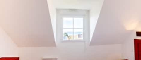 Um quarto com teto inclinado, janela com vista, cama, mesa de cabeceira e cômoda vermelha.