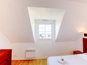 Una habitación con techo inclinado, ventana con vistas, cama, mesita de noche y cómoda roja.