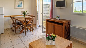 A dining area with a wooden table and chairs, a small wooden cabinet, a TV mounted on the wall, and a window with a view of greenery. - Residence Vacanceole Les Terrasses De Pentrez (Saint-Nic)