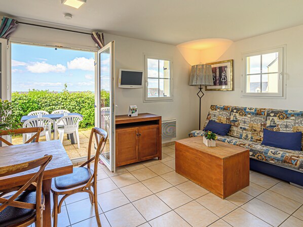 Sala de estar con sofá, mesa de centro y mesa de comedor con sillas. Hay un televisor y una ventana con vista a zonas verdes.