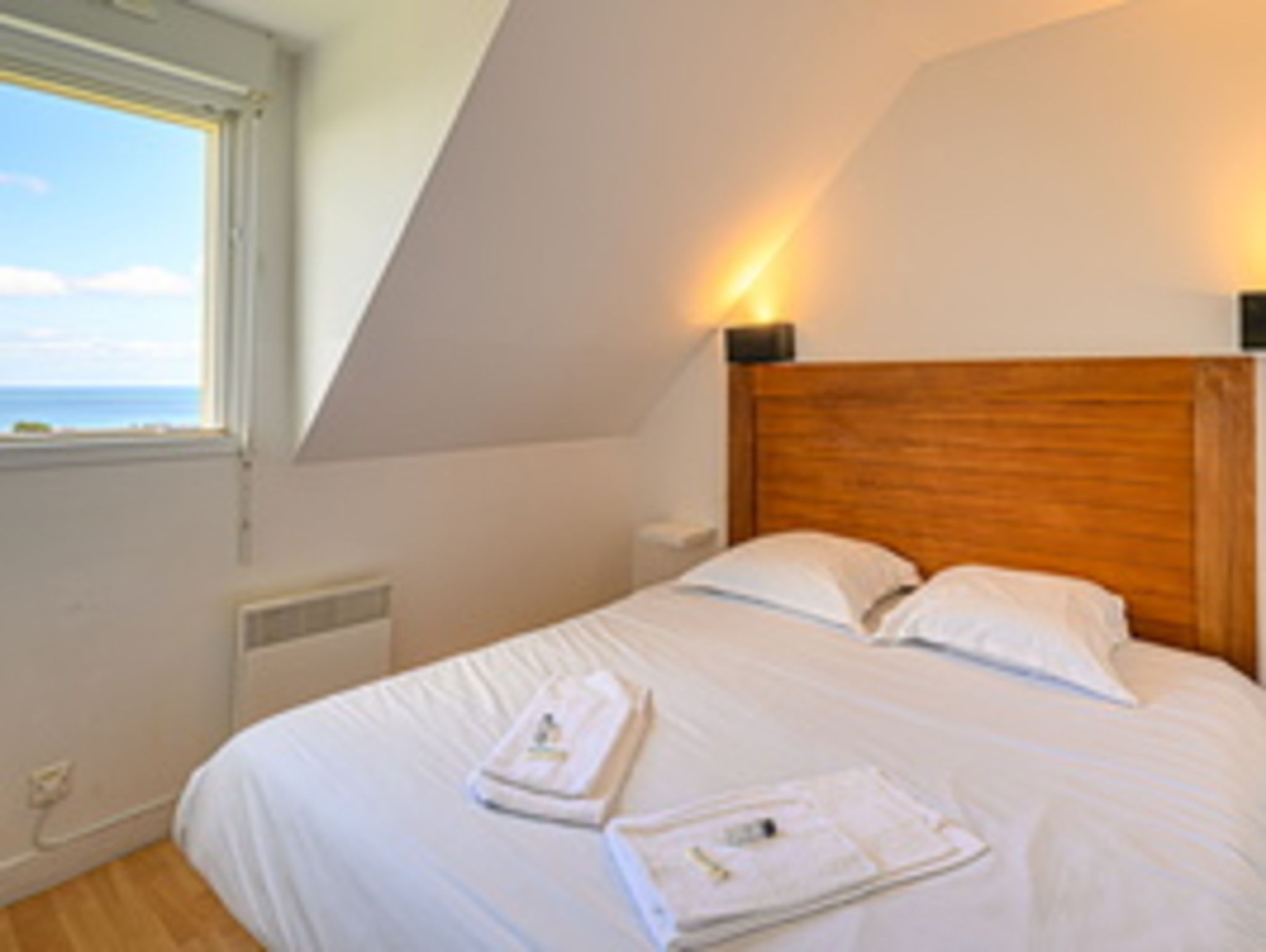 A bedroom with a sloped ceiling, a wooden headboard, white bedding, and a view of the sea through a window.