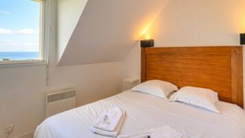 A bedroom with a sloped ceiling, a wooden headboard, white bedding, and a view of the sea through a window.