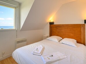 Ett sovrum med ett sluttande tak, en sänggavel i trä, vita sängkläder och utsikt över havet genom ett fönster.