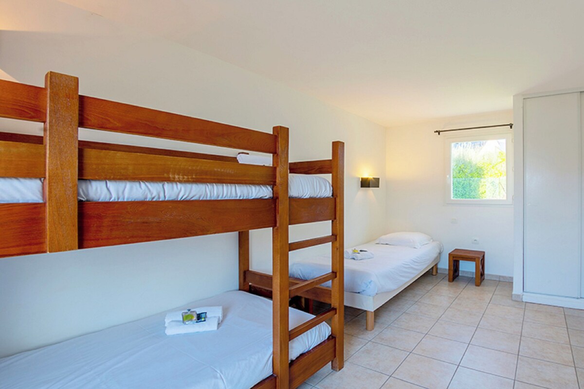 A room with two bunk beds and a single bed, a small bedside table, and a window with a view of greenery.