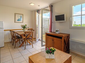 Un comedor con mesa y sillas de madera, un mueble de madera pequeño, un televisor montado en la pared y una ventana con vistas a zonas verdes.