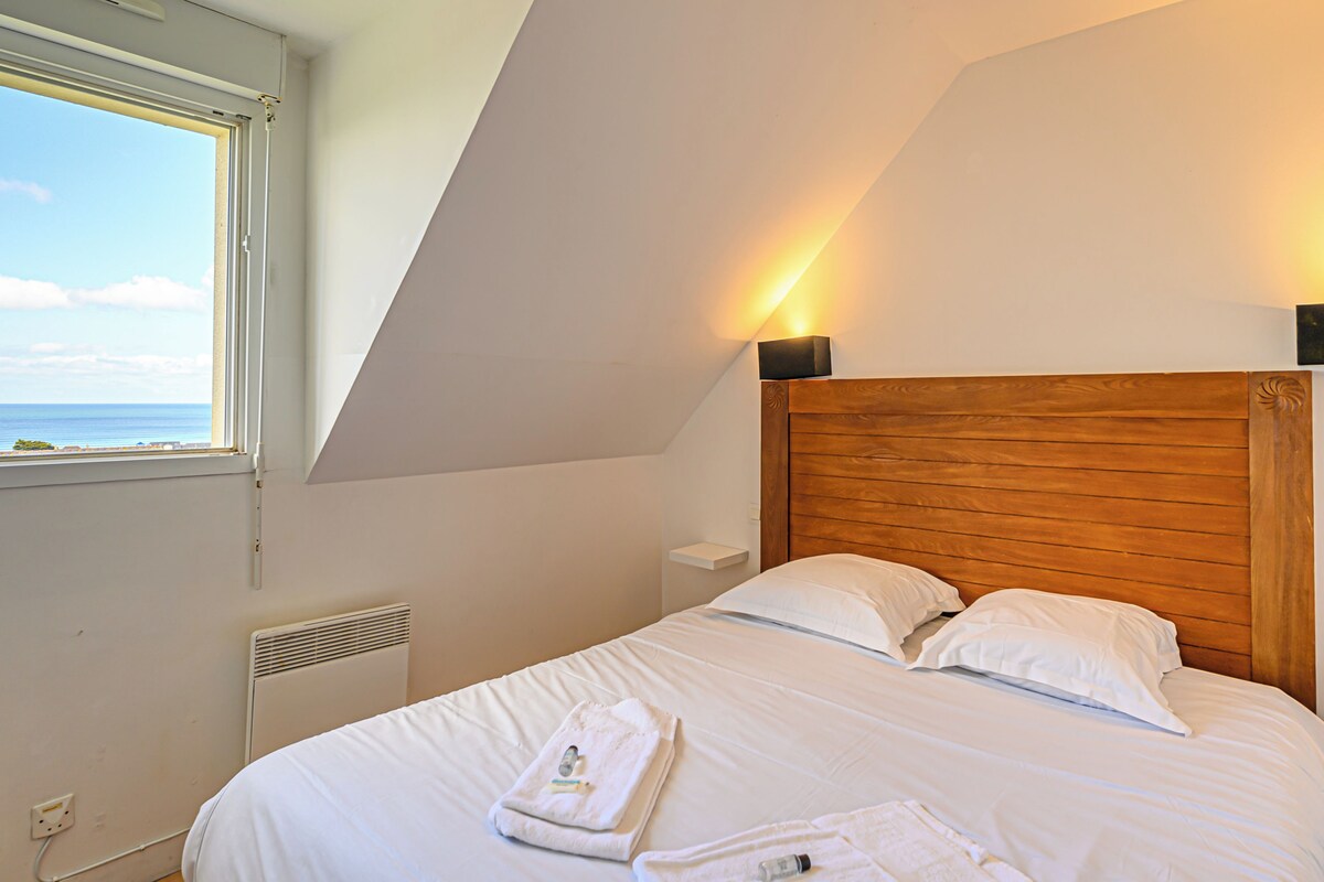 Un dormitorio con cabecera de madera, cama con ropa de cama blanca y vistas al mar a través de una ventana.