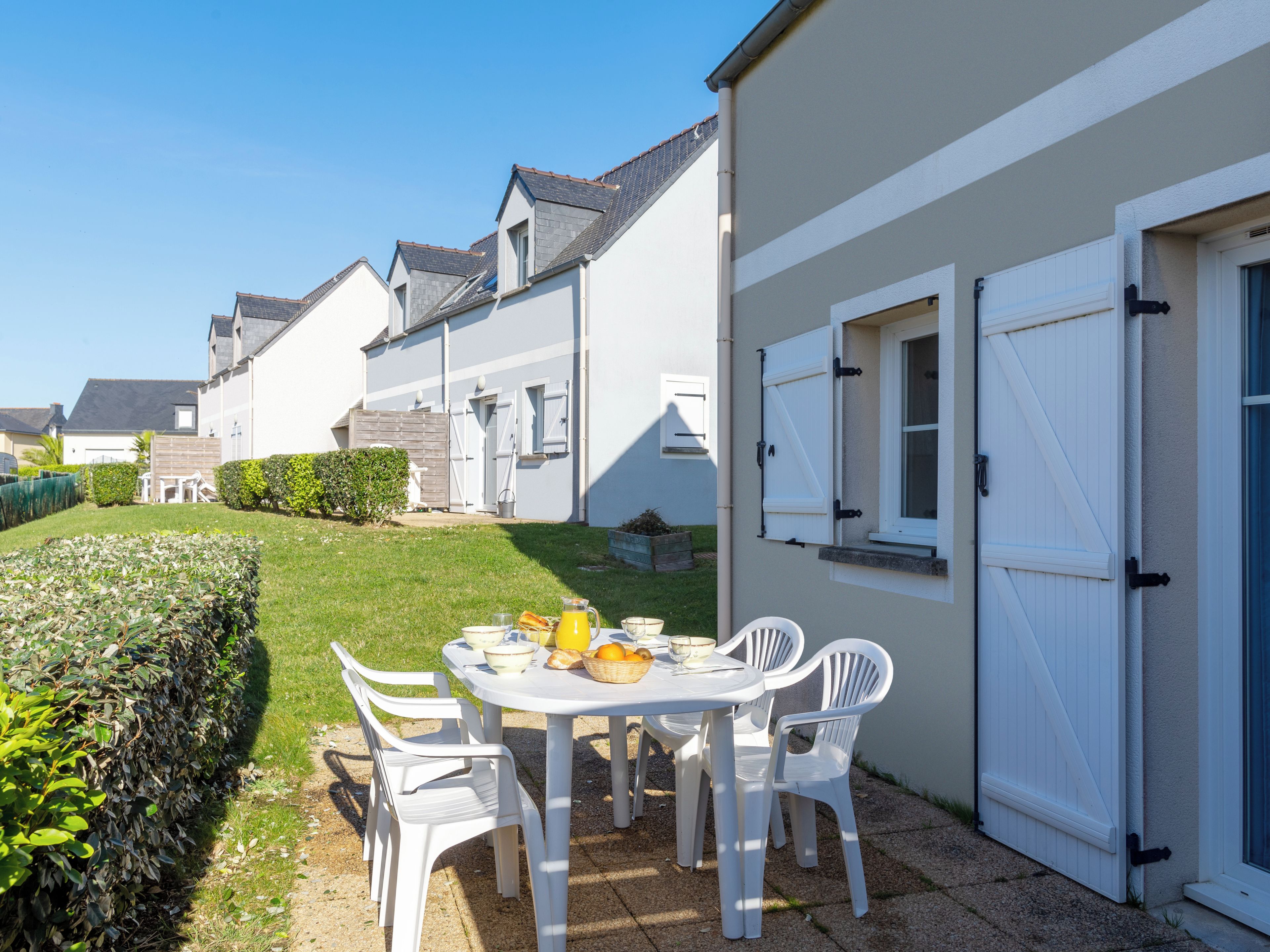 Un patio con una mesa lista para el desayuno, rodeado de sillas blancas, en una zona residencial con casas.