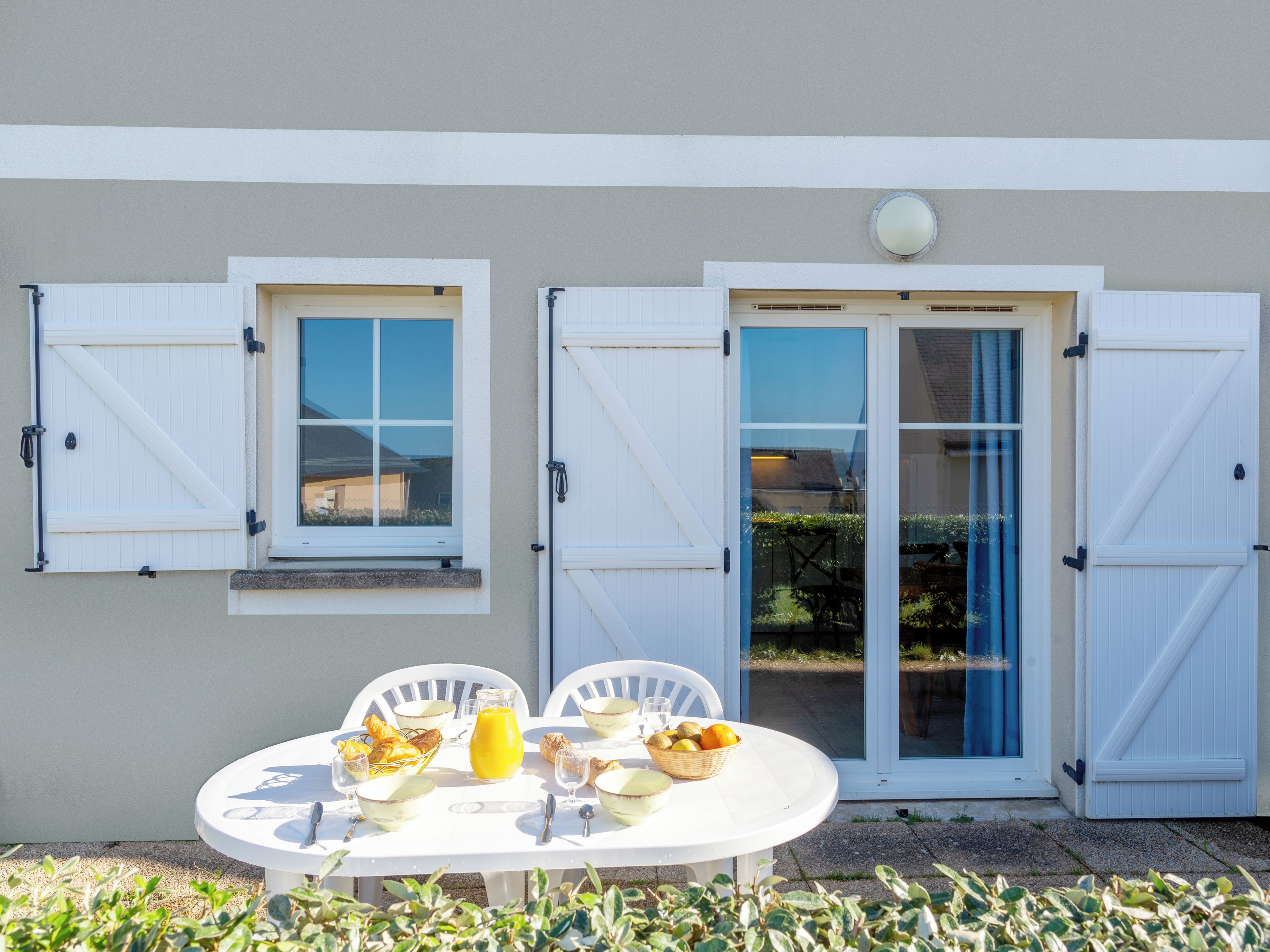 A dining table set outdoors with a view of a house, featuring a window, a door, and a small balcony.