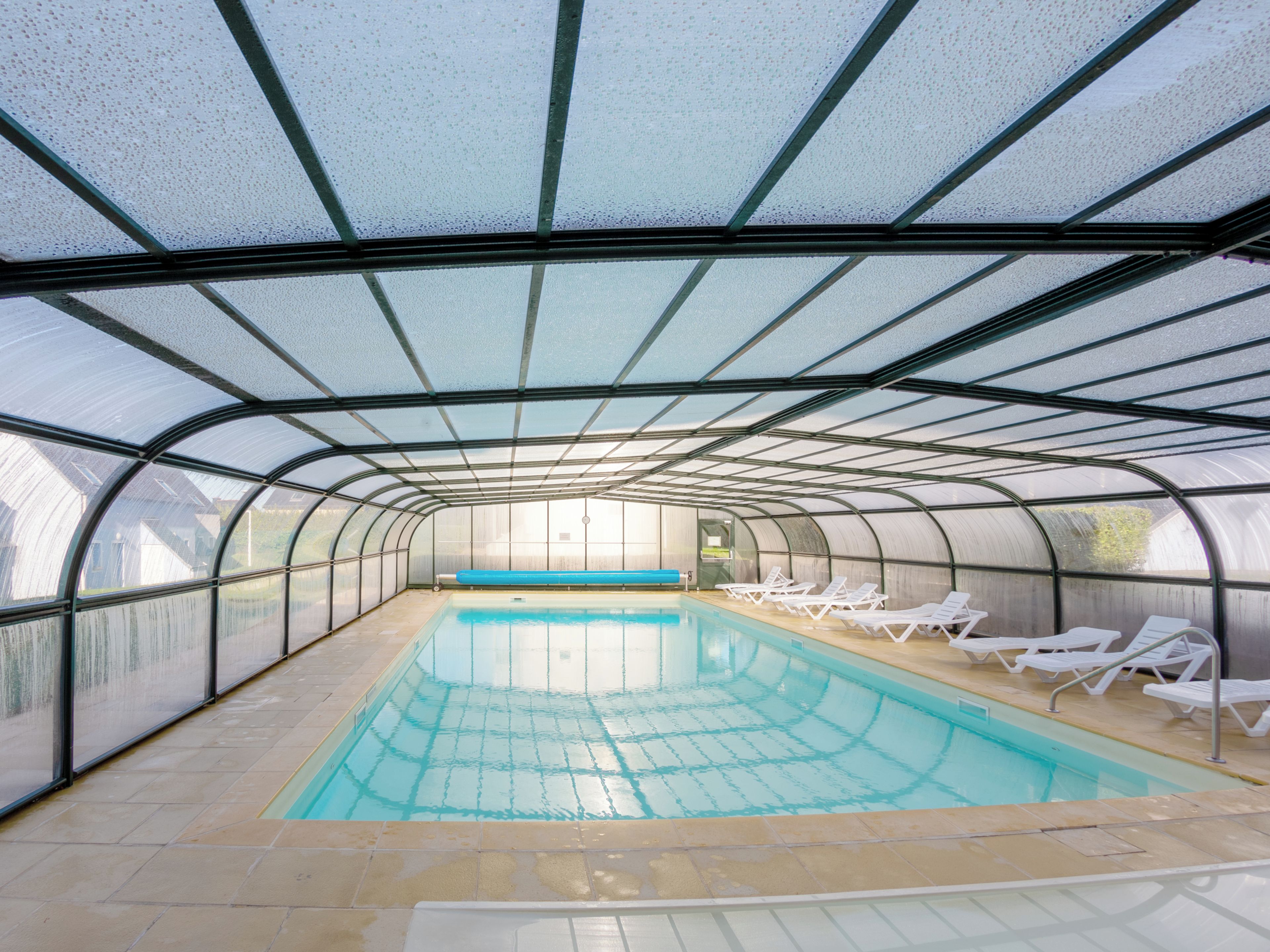A covered swimming pool with clear water, surrounded by white lounge chairs and a glass ceiling structure.