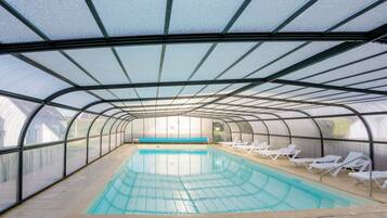 A covered swimming pool with clear water, surrounded by white lounge chairs and a glass ceiling structure.
