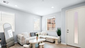 Un salon moderne avec un canapé beige, une table basse blanche et une petite table d’appoint. Il y a deux fenêtres avec des stores, une porte et une plante en pot.