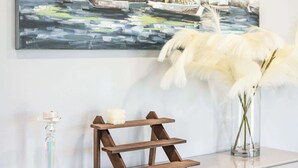 Un tableau représentant des voiliers sur l’eau, un vase avec des fleurs blanches et une étagère en bois posée sur une commode blanche.