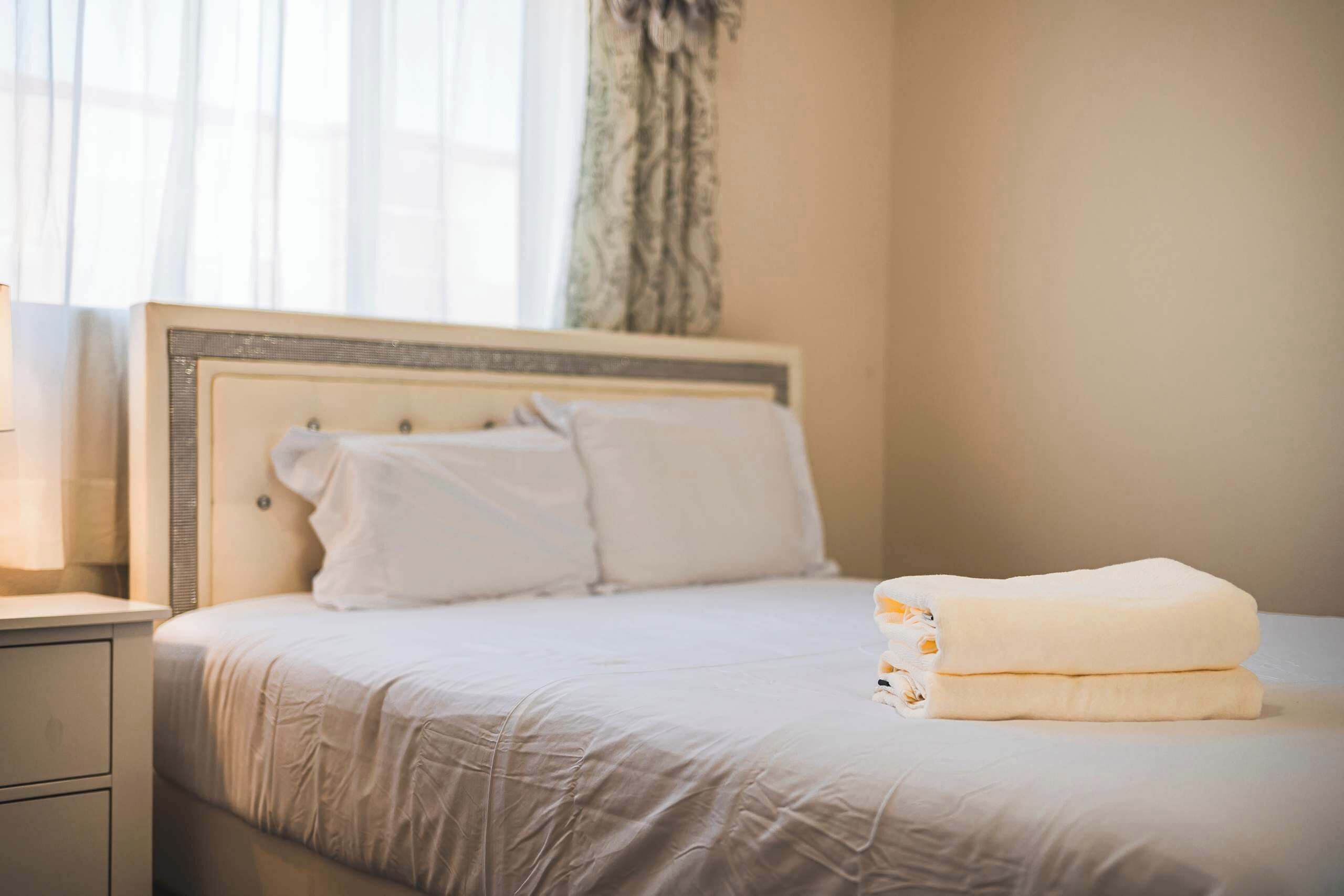 Una cama bien hecha con sábanas blancas, una mesita de noche y una ventana con cortinas traslúcidas.