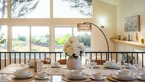Une table à manger dressée avec de l’assiette, des bols et des verres blancs, un vase rempli de fleurs blanches, et une vue sur la verdure à travers de grandes fenêtres.