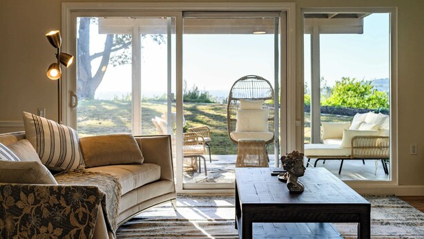 Un salon comprenant un canapé, une table basse et des fauteuils en osier. Une grande porte-fenêtre coulissante donne accès à un espace extérieur avec vue sur la verdure.